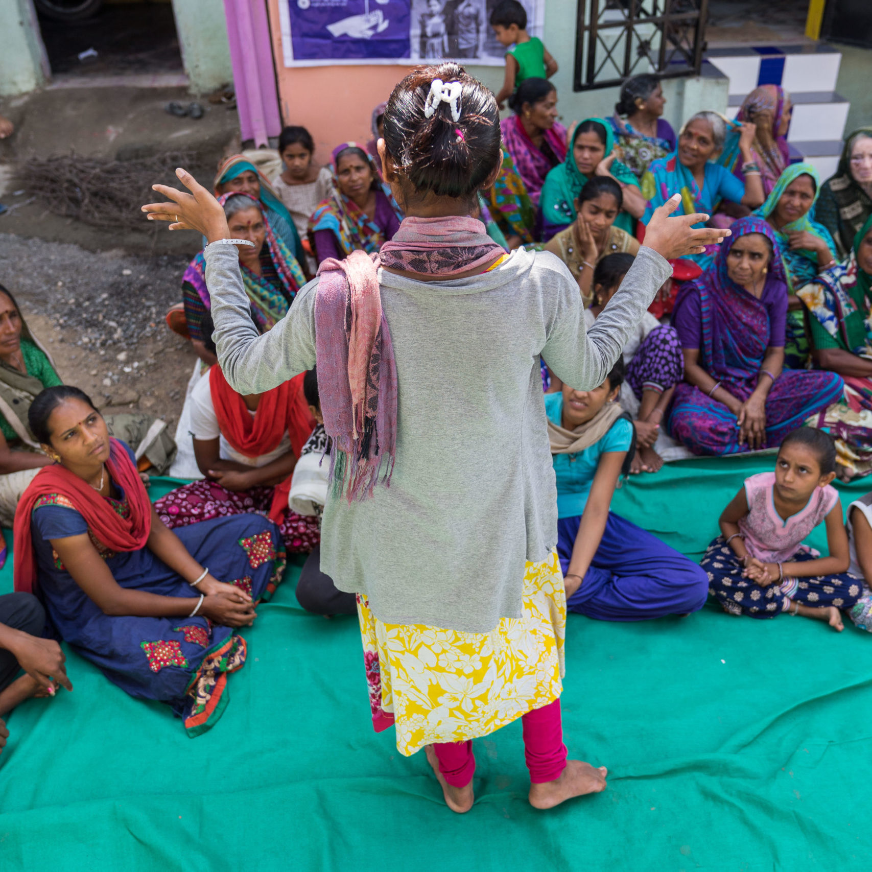 A woman presents to a seated group of women outdoors in India.
