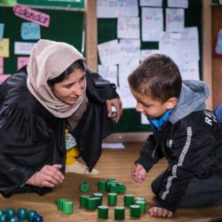 a female teacher in a headscarf sits next to a boy student on the ground. They are using wooden blocks as a hands-on teaching tool in a well-appointed classroom