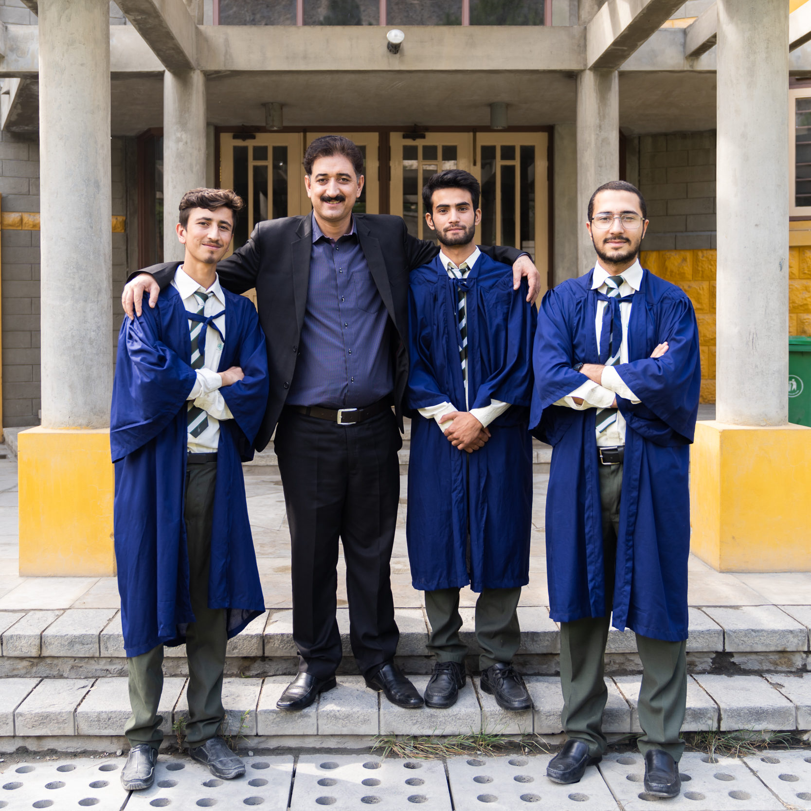 three young men wearing graduation gowns stand with a teacher outside a modern school building