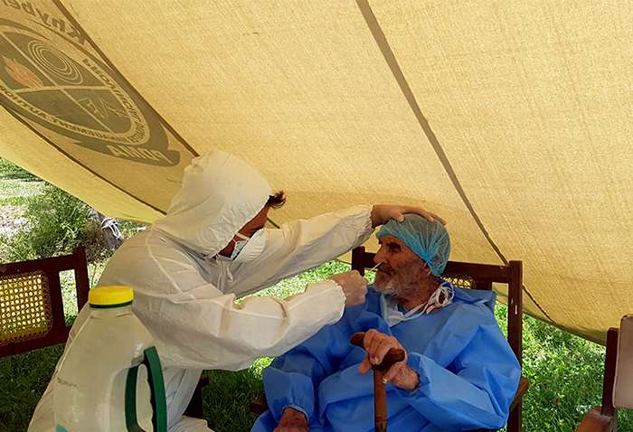 A health professional performs a nasal swab on an elderly man sitting and holding a cane. In the background is a yellow tent background and grass underneath.