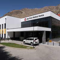 The front of a hospital building with an SUV in front and mountains in the background. "Aga Khan Medical Centre" is on the top of the hospital.