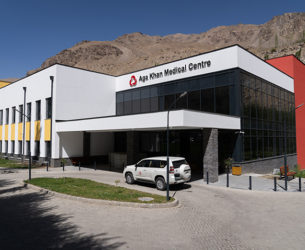 The front of a hospital building with an SUV in front and mountains in the background. "Aga Khan Medical Centre" is on the top of the hospital.