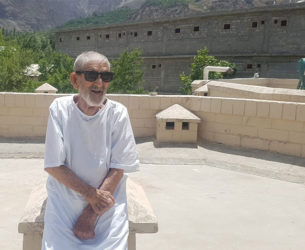 An elderly man wearing sunglasses sits on a rooftop in front of mountains