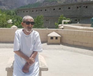 An elderly man wearing sunglasses sits on a rooftop in front of mountains