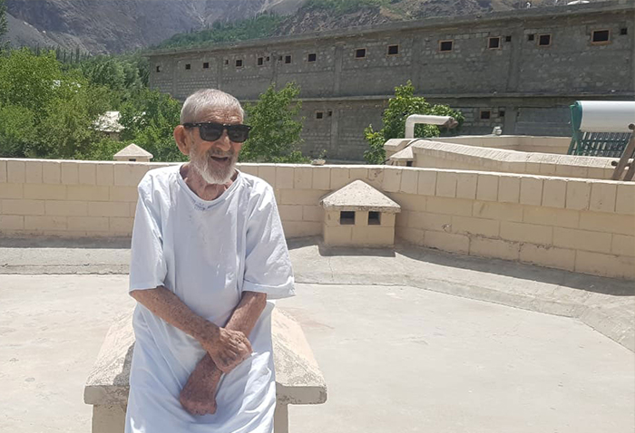 An elderly man wearing sunglasses sits on a rooftop in front of mountains