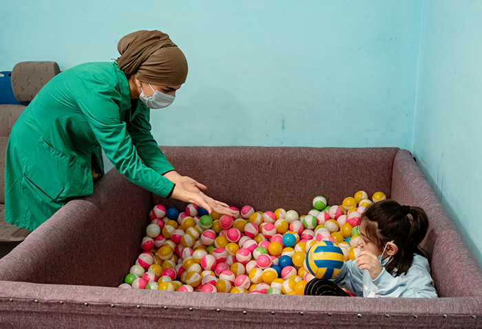 A young girl sits in a ball pit holding a ball; a teacher standing outside the ball pit reaches out to her.