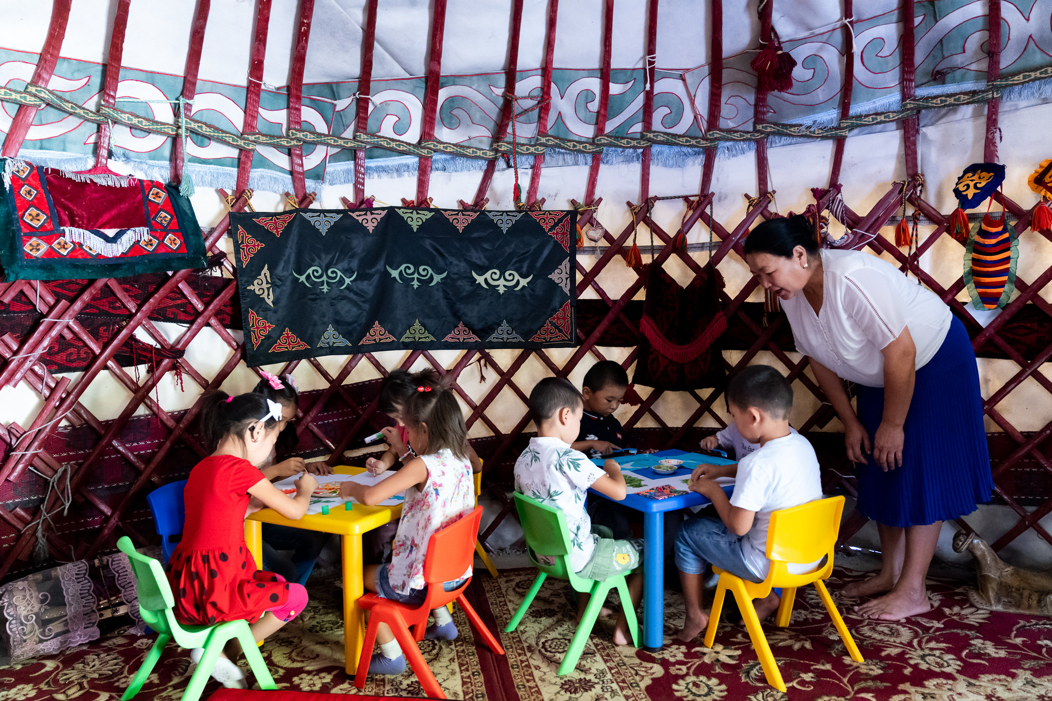 Four young children sit at two colorful desks as a teacher leans over to look at their work