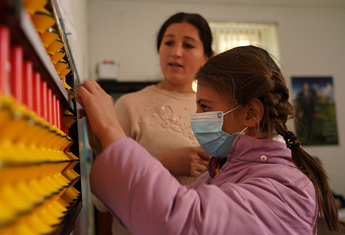 A young girl wearing a mask stands by an abacus. A teacher stands behind her providing instructions.