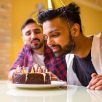 A man blows out candles on a cake with a friend in the background.