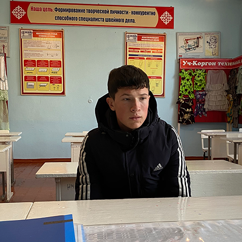 A young man sits on a chair with am empty classroom behind him.