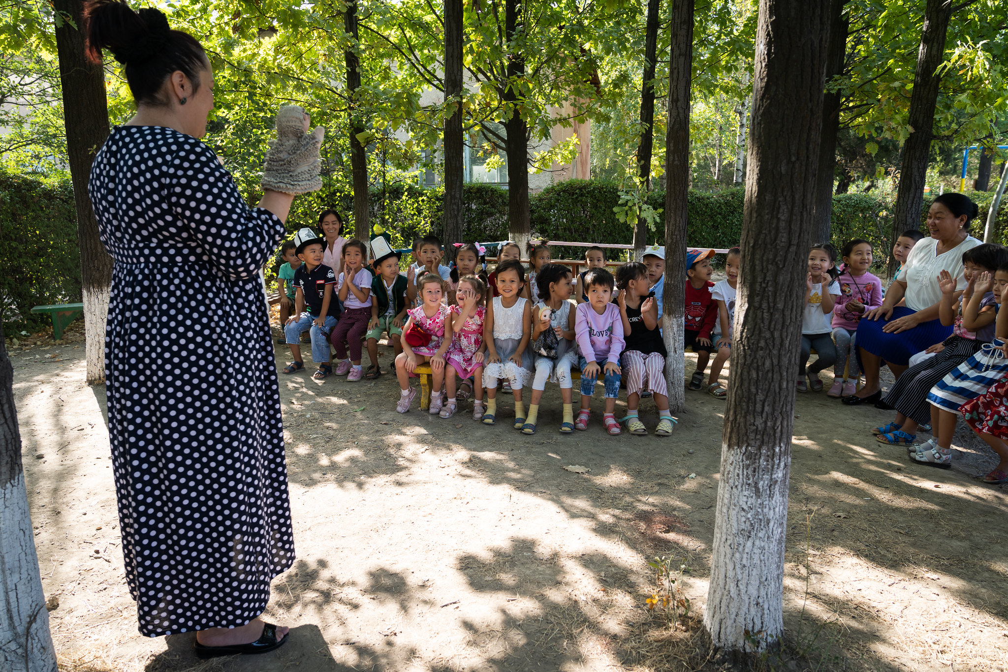 A teacher stands outside with a hand pupper as rows of young chidlren sit on benches watching her