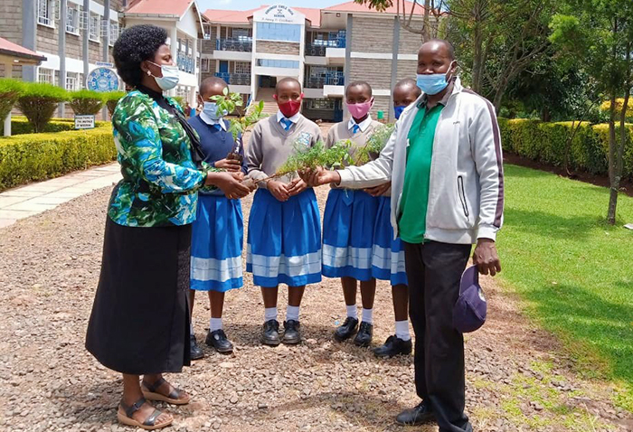 Three girls in school uniforms hold seedlings in the middle of a school official and a representative from Transformers CBO. They are standing in front of the school buildings.