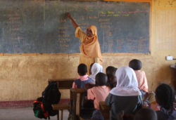 A teacher points to words on a chalkboard in front of a classroom of students.