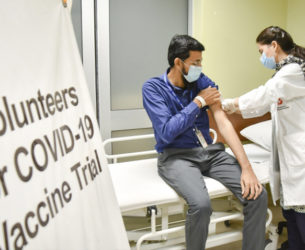 A doctor adminiters a vaccine to a man sitting on a hospital bed next to a banner that says "Volunteers for COVID-19 Vaccine Trial"